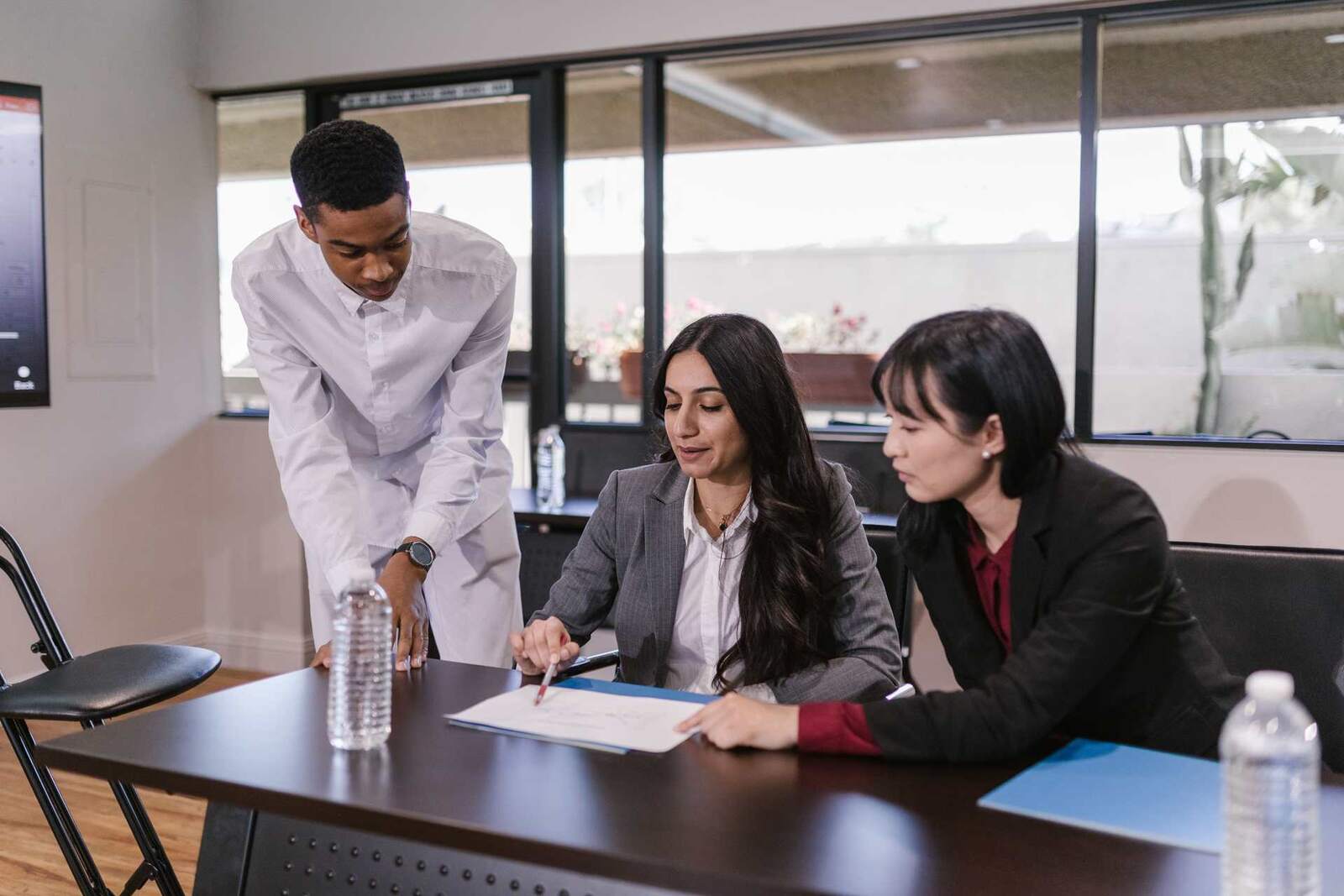 A colleague leans over a table to point at a document while two seated colleagues review it in a bright meeting room