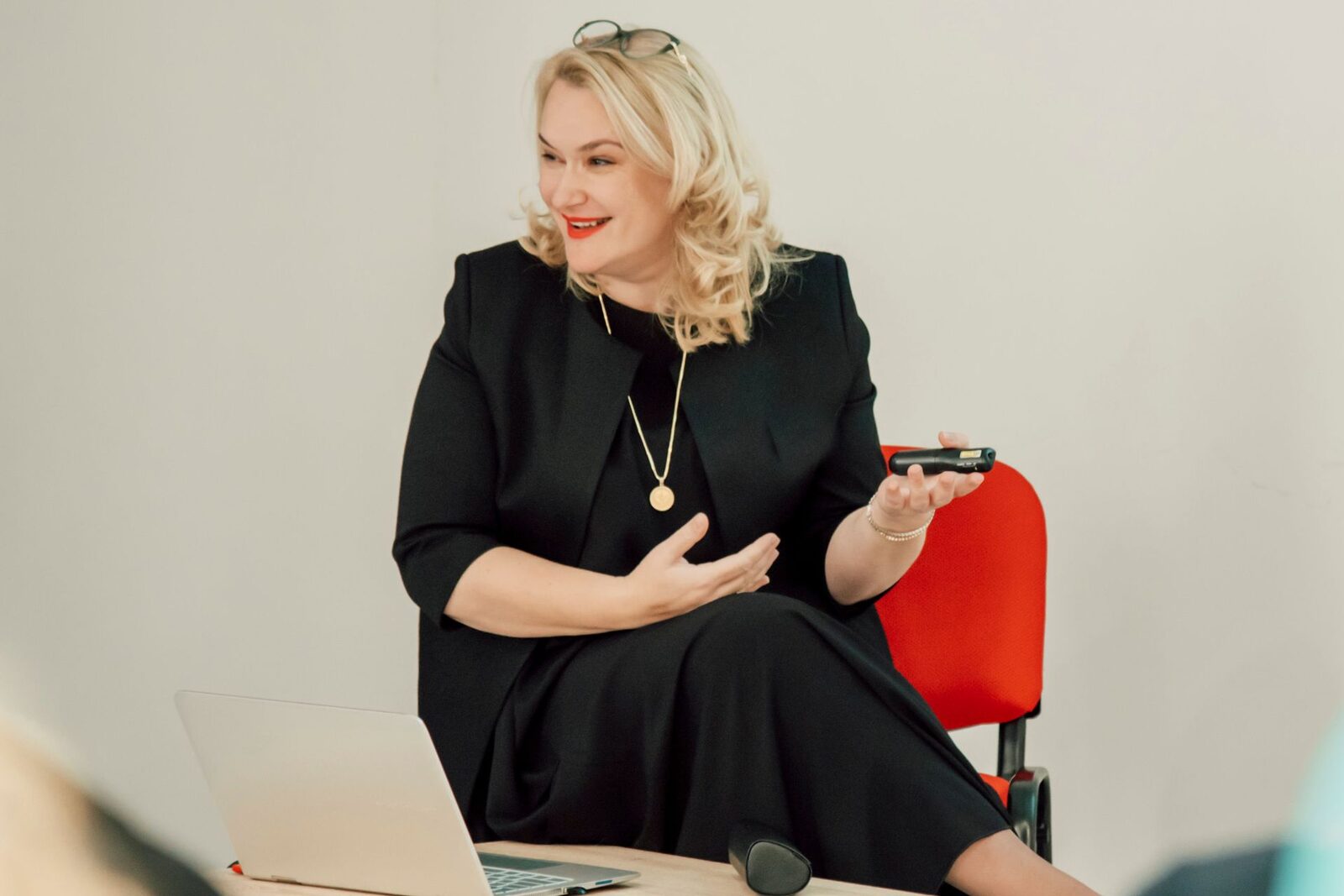 A smiling woman speaker in a black dress and jacket sits beside an open laptop holding a slide clicker, bright even light, in a plain workshop room