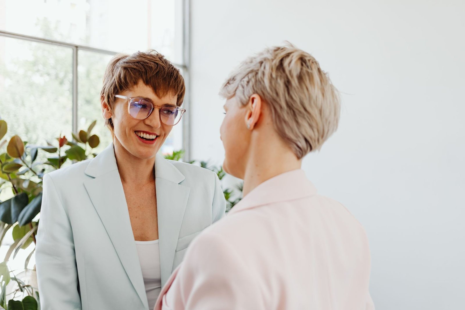 Two women in pale blazers laugh mid-conversation beside a bright window and potted plants