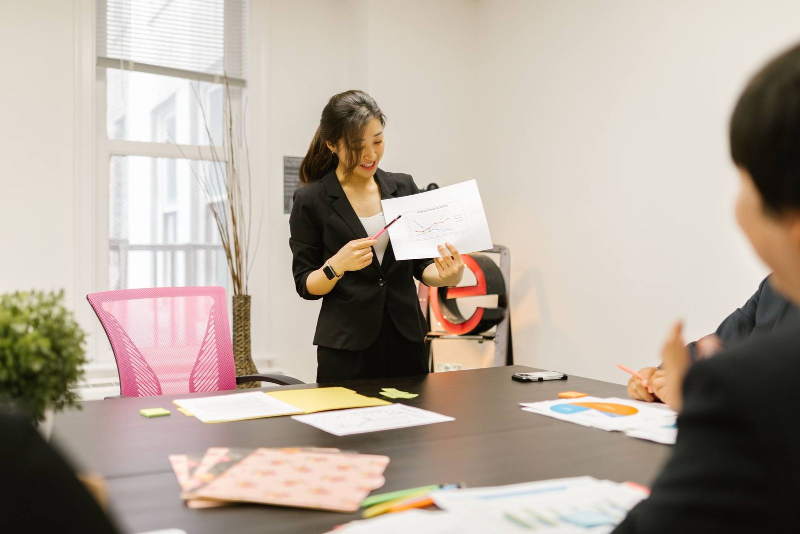 A woman in a black blazer smiles while holding up a printed line chart labeled Product Trends by Month, pointing to it with a pen for a seated colleague in a bright meeting room