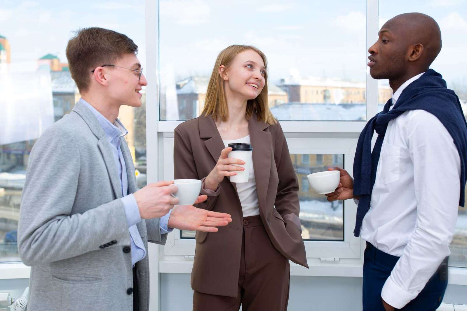 Three colleagues in business dress talk easily over coffee cups beside a tall bright window