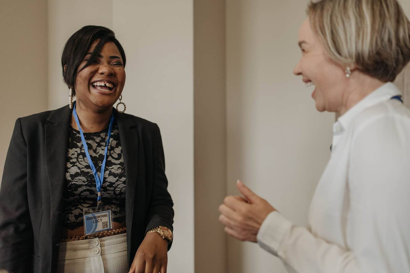 Two women with conference lanyards laugh together mid-conversation, one wearing a visible 