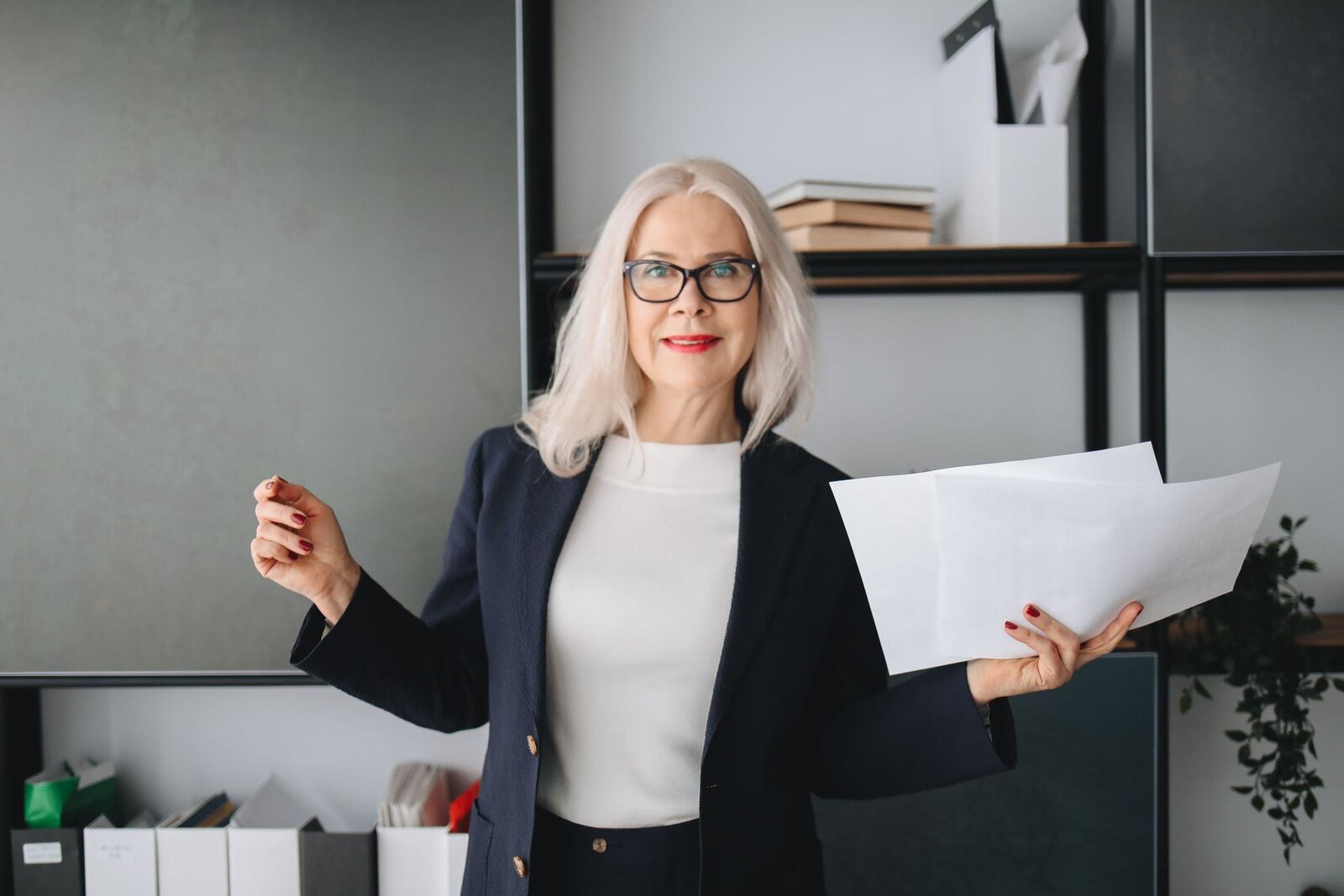 A woman in a navy blazer holds a sheaf of papers mid-gesture in a bright office lined with shelves