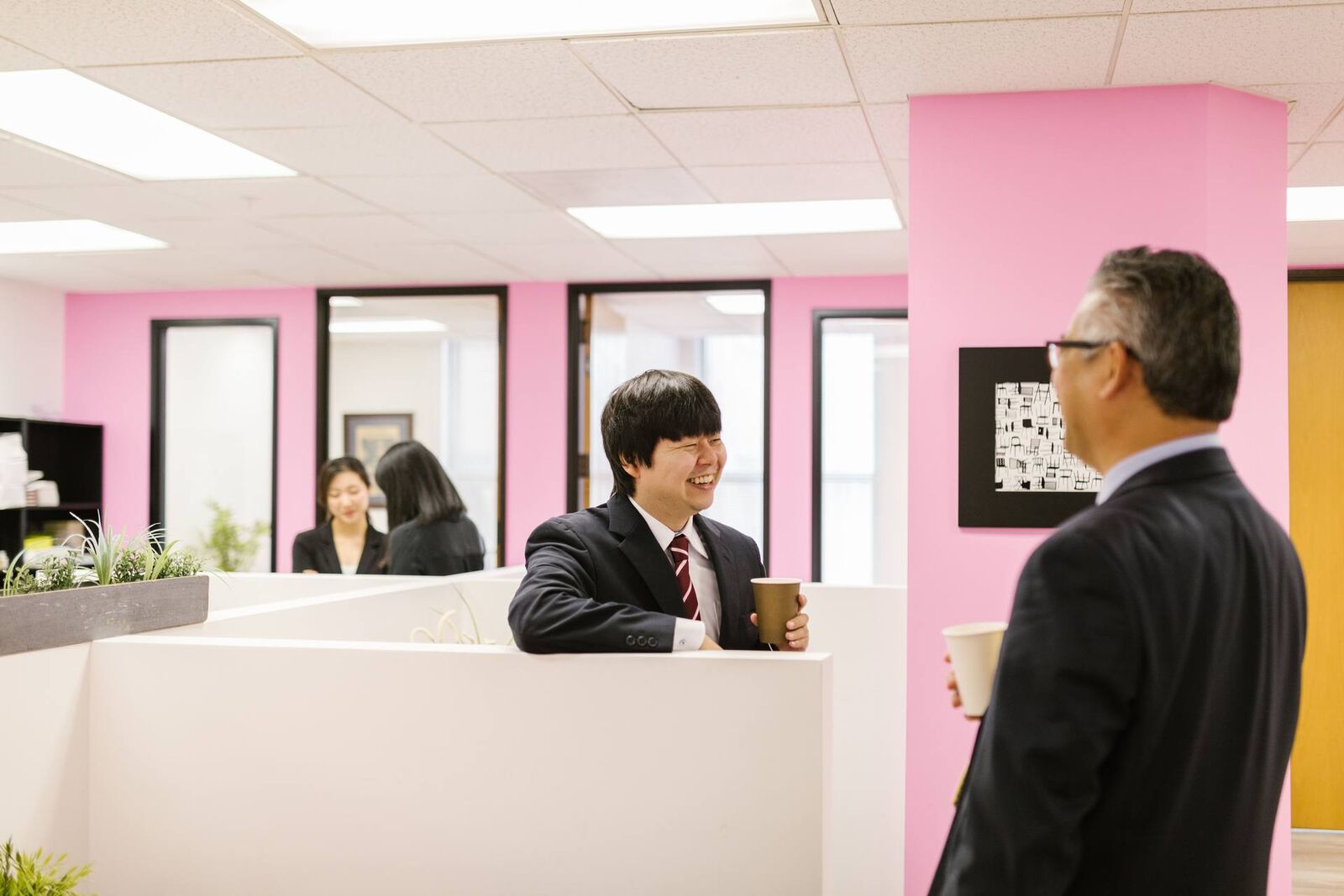 A man in a suit and tie leans over an office partition, caught mid-laugh in conversation with a senior colleague holding a coffee cup, in a bright pink-walled office