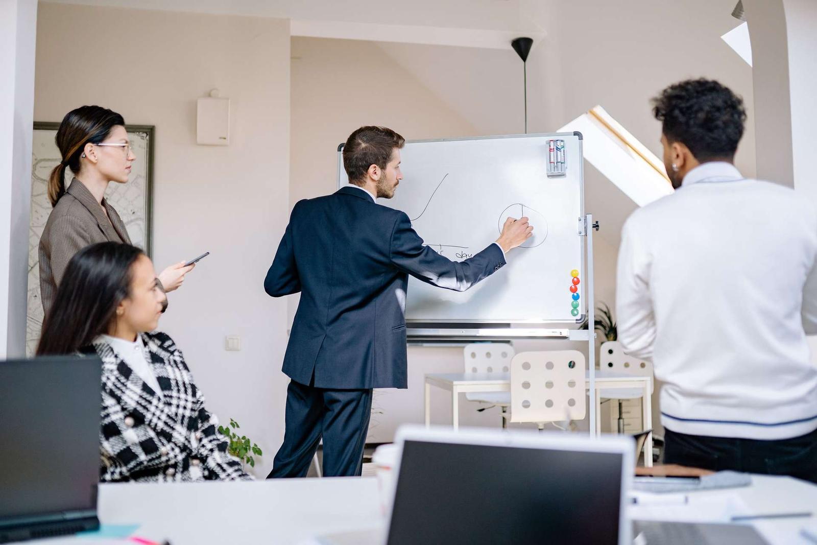 A man in a navy suit stands at a whiteboard sketching a diagram for three colleagues watching from a bright meeting room, laptops open on the table in front of them