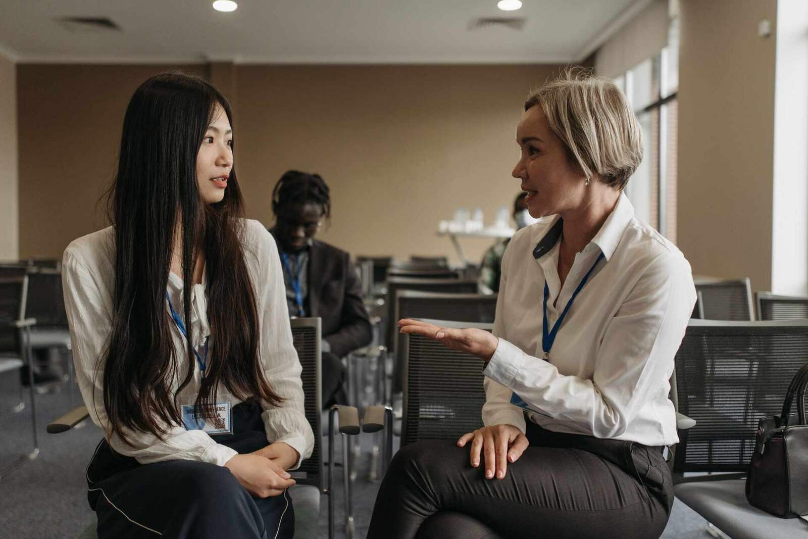 Two women with conference badges talk seated between rows of chairs, window light at right