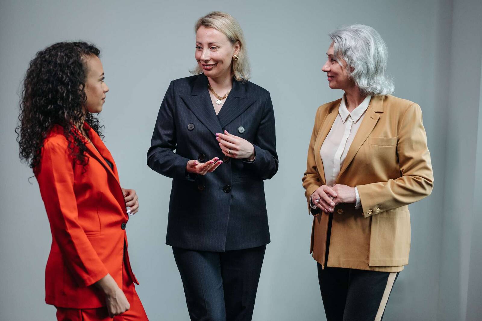 Three women in colorful blazers — orange, navy pinstripe, and camel — stand talking together against a plain grey studio wall