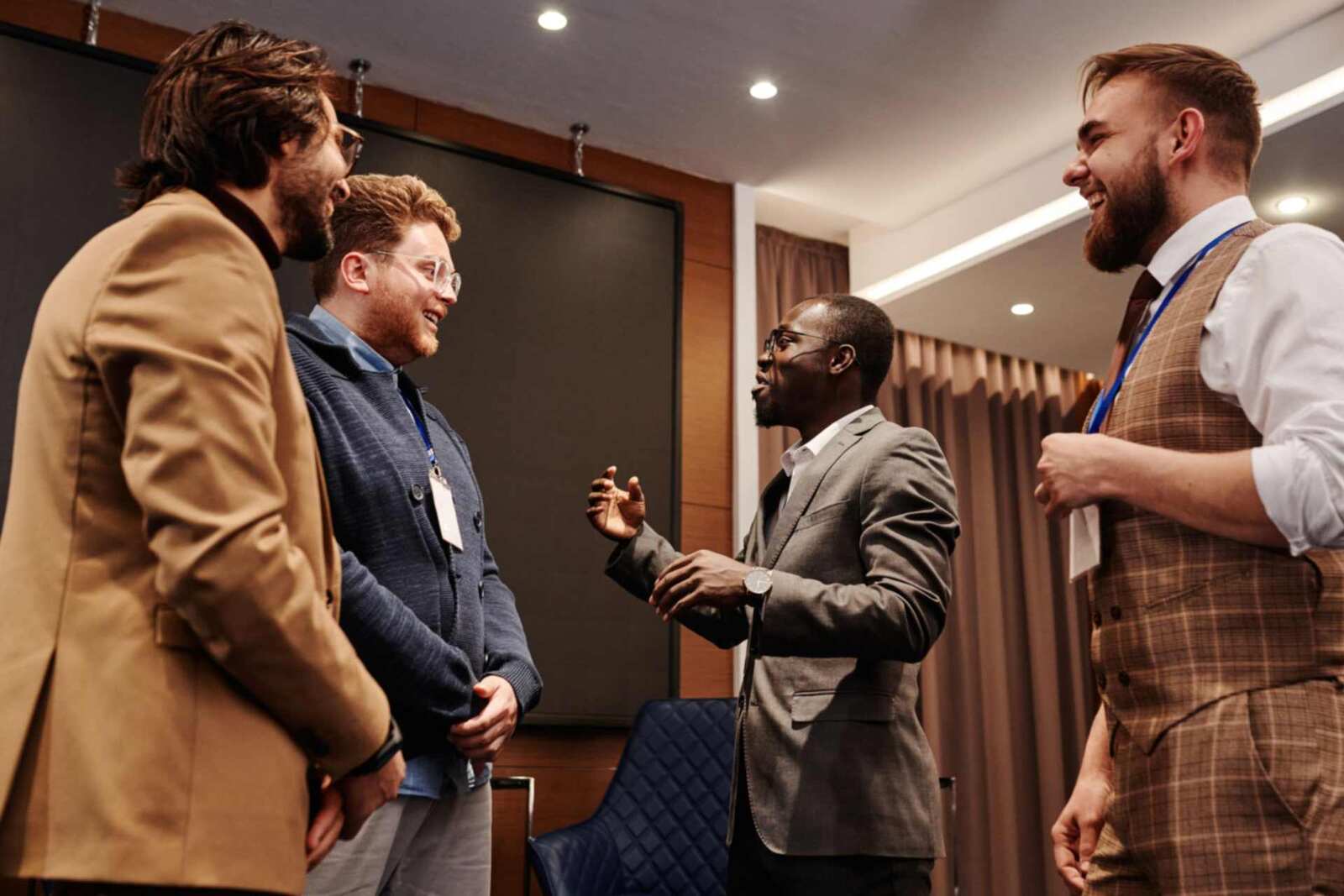 Four men with conference lanyards share a laugh in a hotel meeting suite, warm lighting, a blank screen on the wall behind them