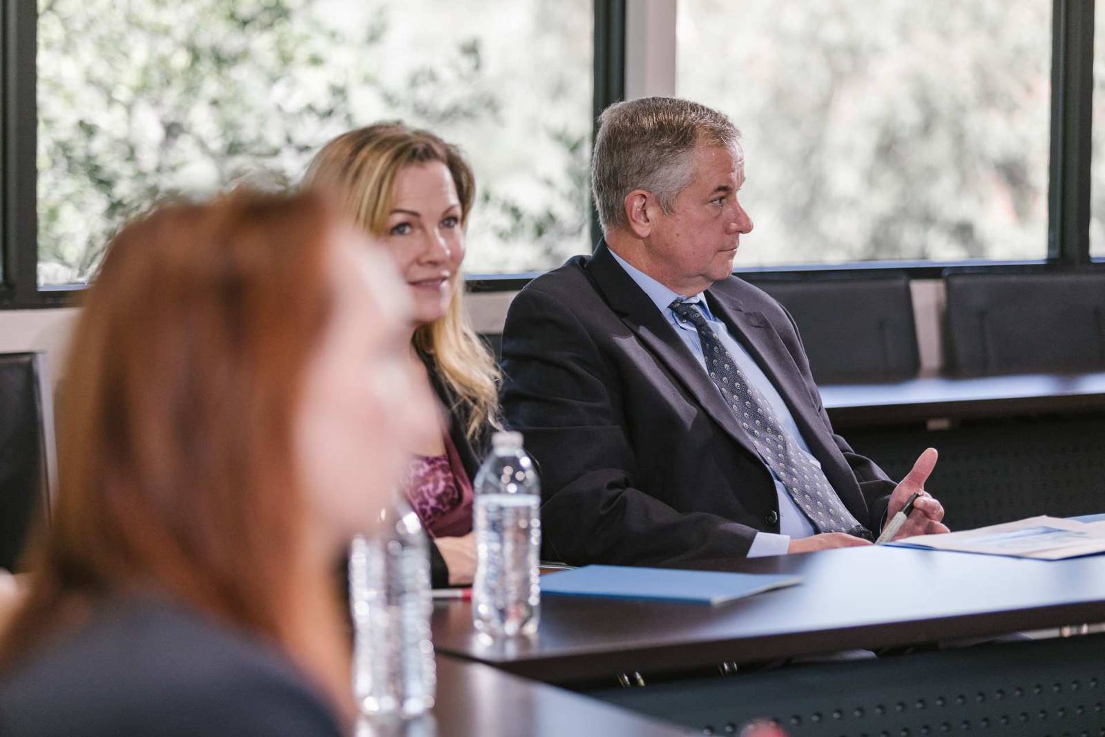 A woman and a man listen attentively at a conference table lined with water bottles, a colleague blurred in the foreground and trees visible through the windows behind them