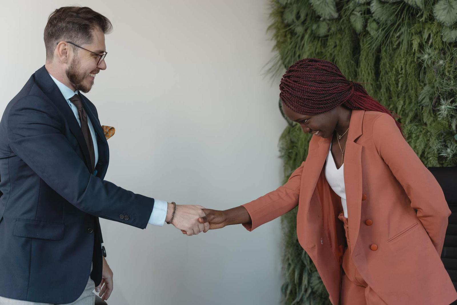 A man in a navy suit and a woman in a coral blazer shake hands and smile, in front of a green plant wall