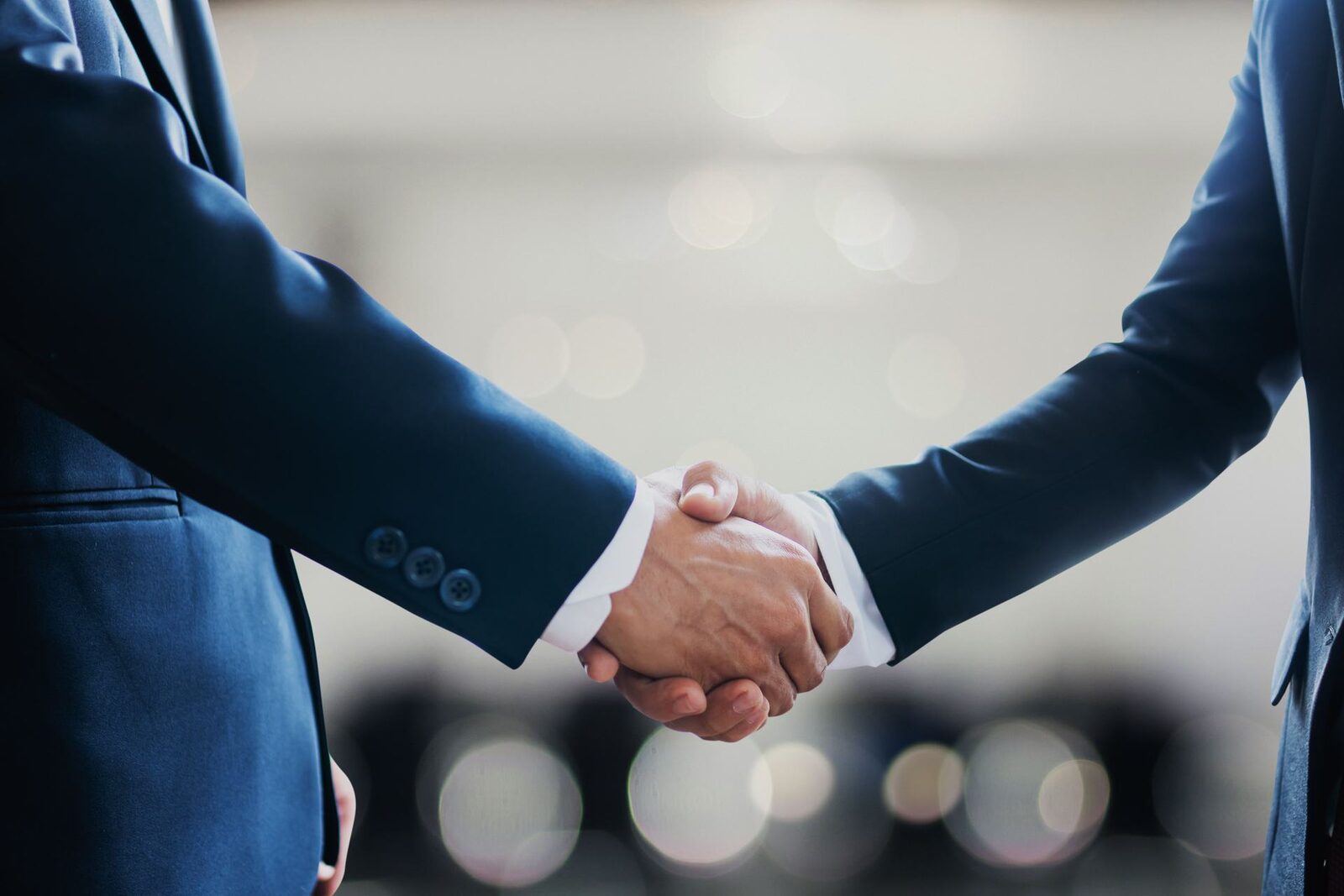 Close-up of two men in navy suits shaking hands, white shirt cuffs visible, against a bright, blurred venue background