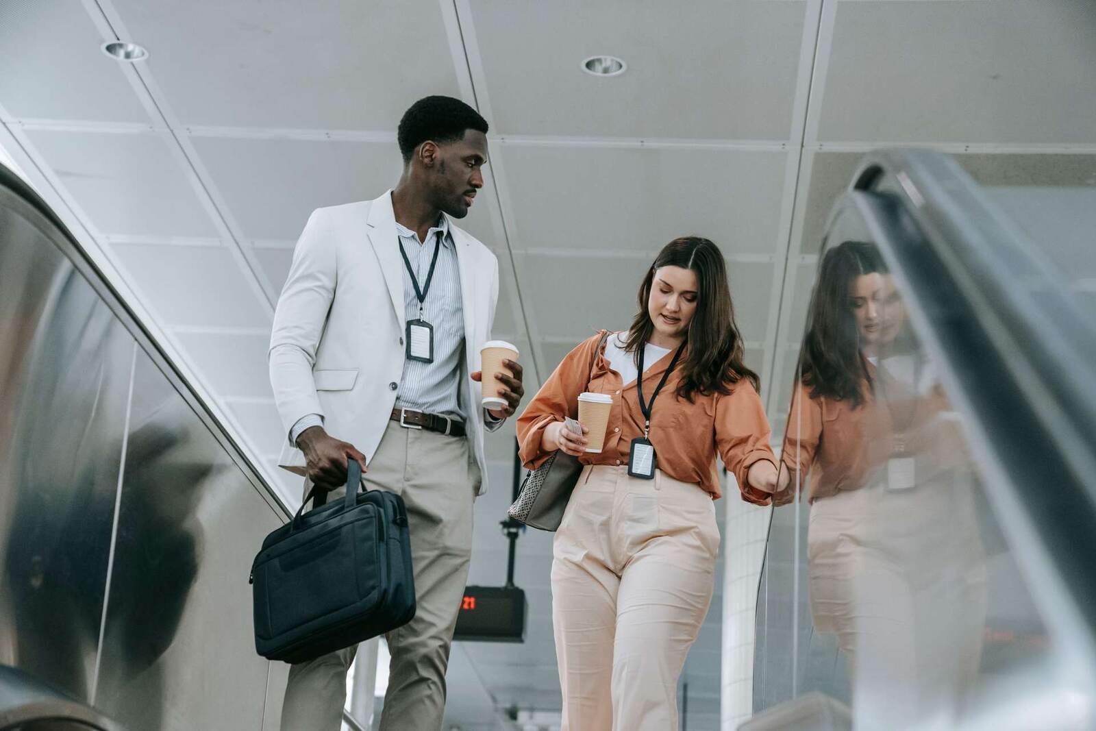 A man with a laptop bag and a woman, both wearing lanyards and holding coffee, step off an escalator together in a bright concourse
