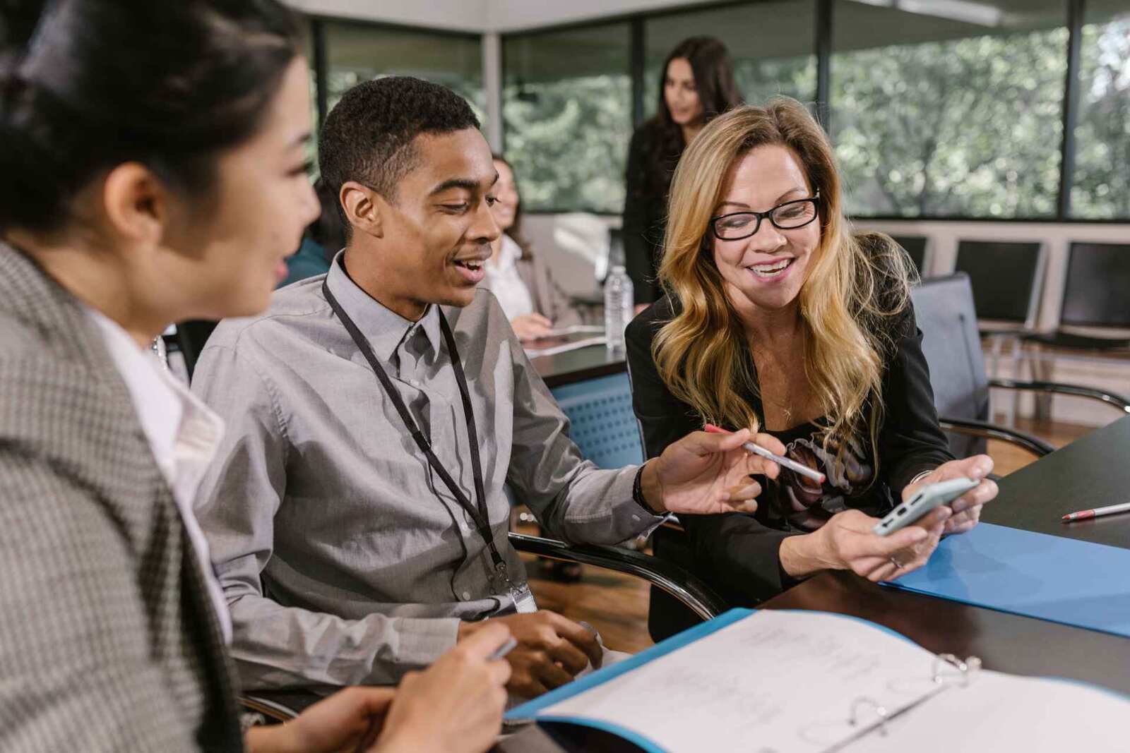 A young man with a conference lanyard and two smiling colleagues look over a binder and a phone at a table in a bright, glass-walled meeting room