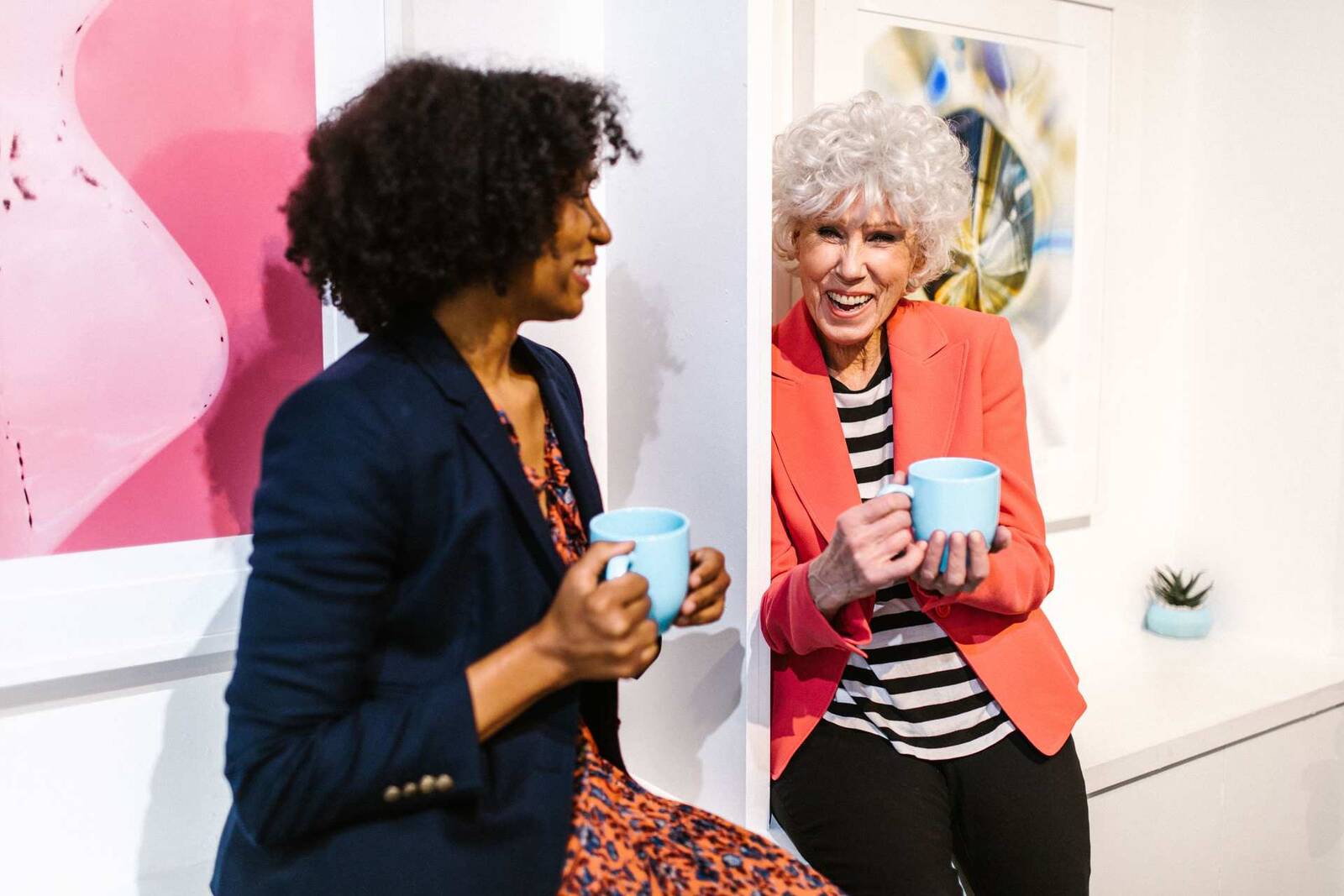 Two women in blazers, one visibly older, laugh together while holding coffee mugs against a colorful gallery wall