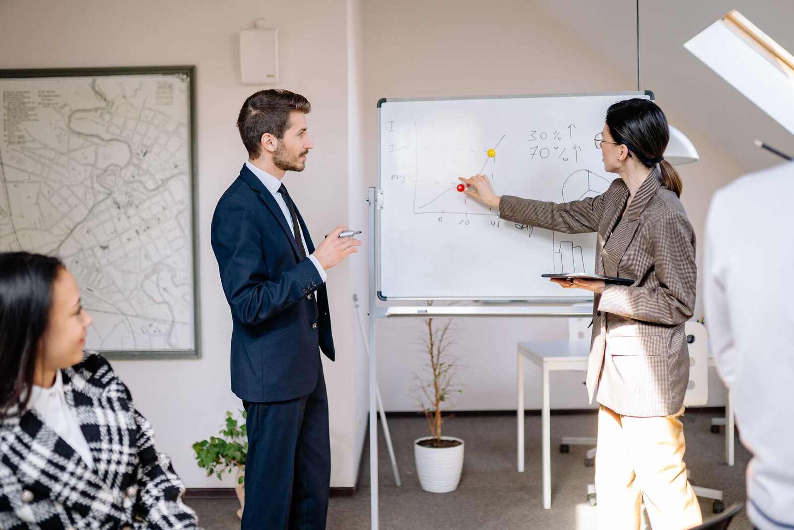 A man in a navy suit and a woman in a houndstooth blazer point to a hand-drawn growth chart on a whiteboard while colleagues look on in the foreground