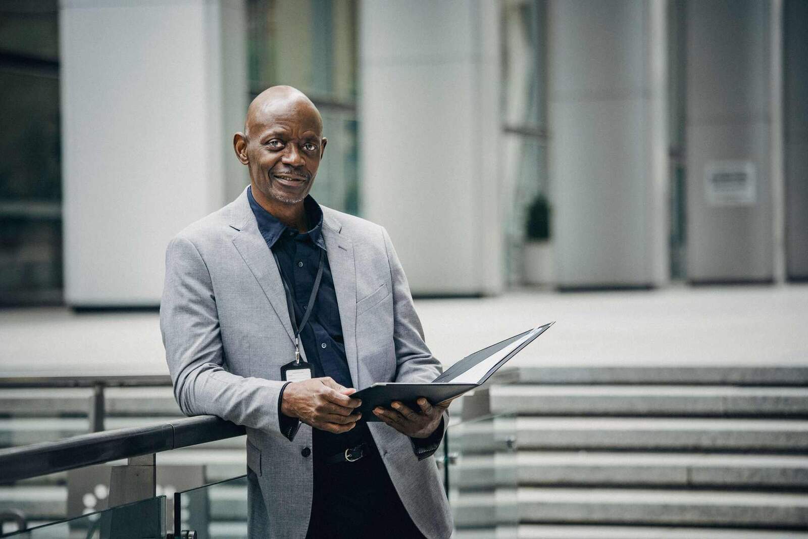 A smiling man in a grey blazer wears a lanyard badge and holds an open folder, leaning against a railing on an office plaza
