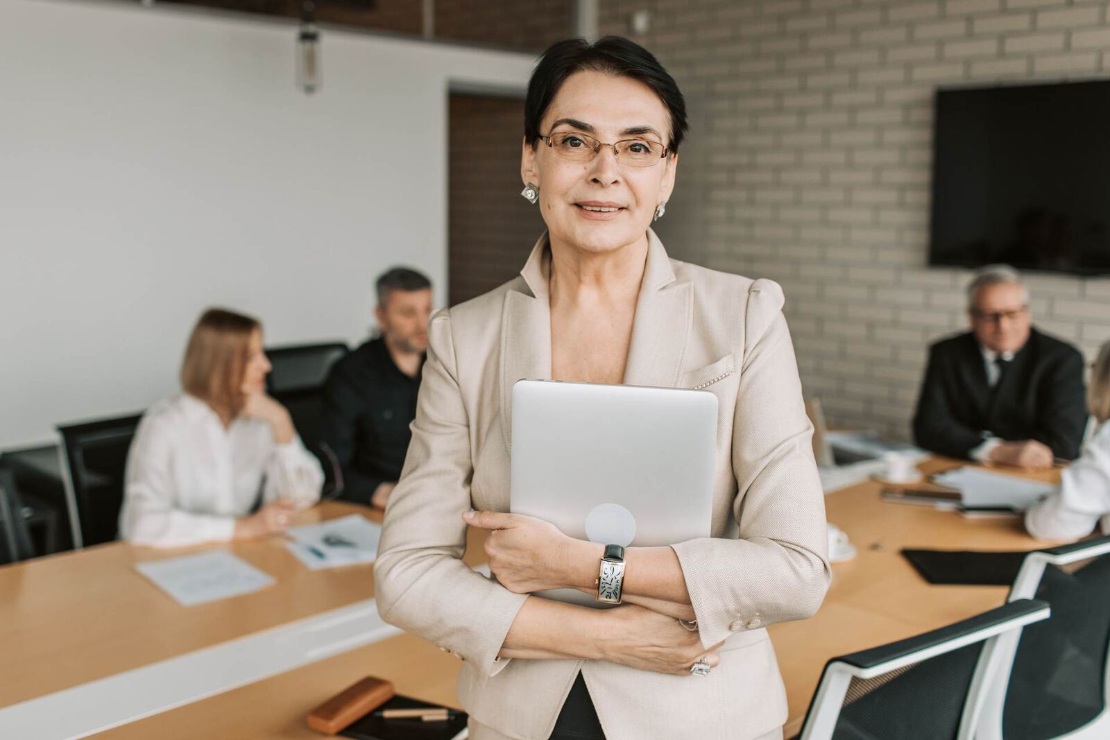 A senior colleague stands holding a laptop to her chest in a bright boardroom, colleagues seated at the table behind her