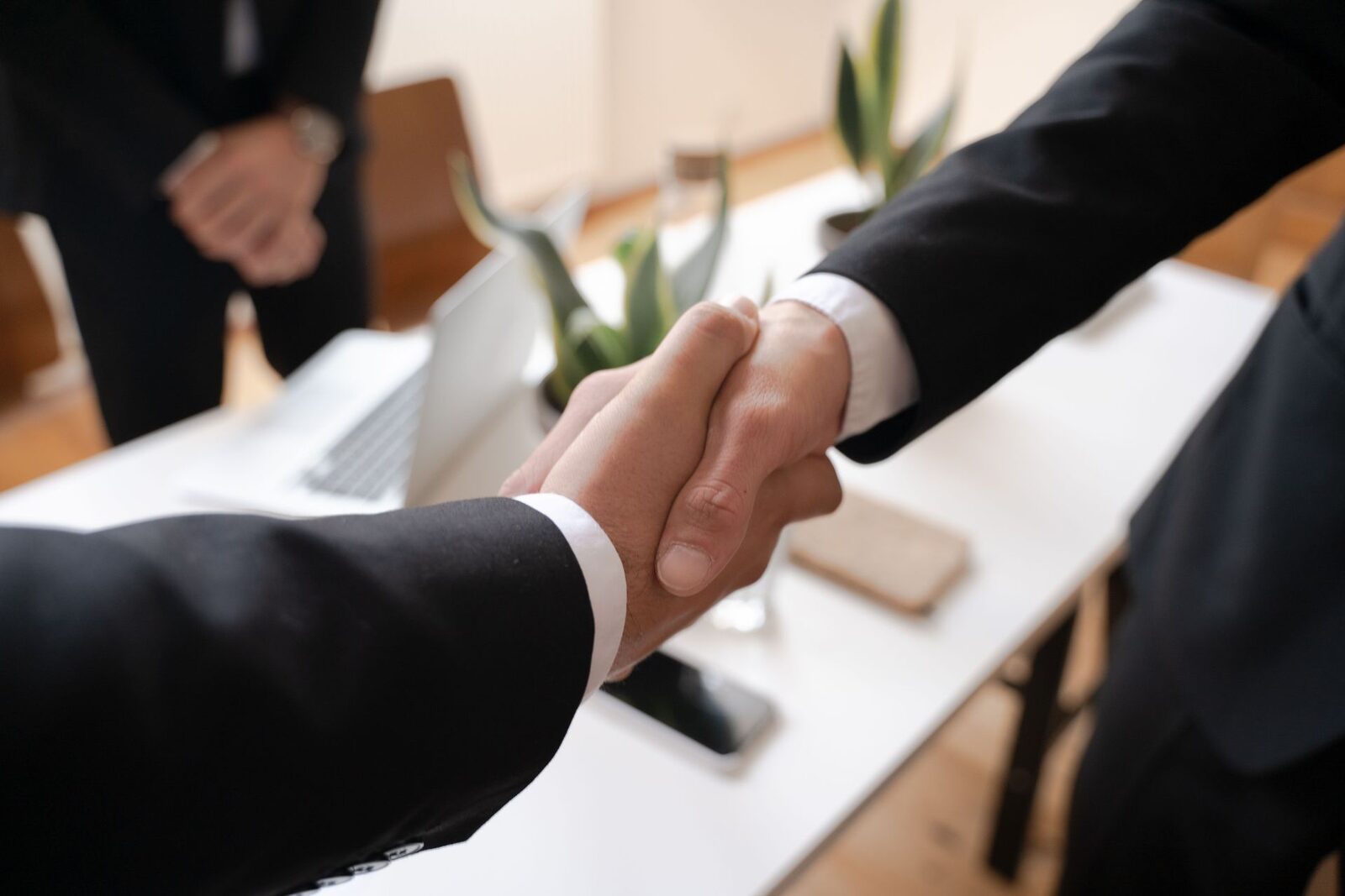 Close-up of two hands shaking, both in black suits with white cuffs, over a white table with a laptop and a plant, a third suited person blurred behind