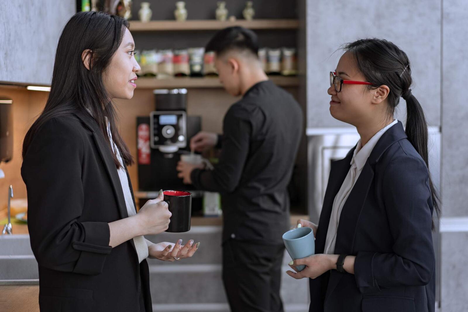 Two colleagues in dark blazers stand talking over coffee mugs at a bright coffee station