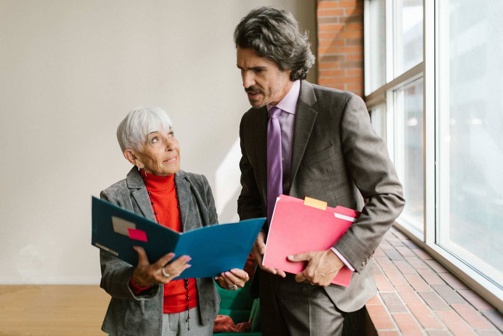 Two senior colleagues in business attire compare open folders of documents in a window-lit corridor