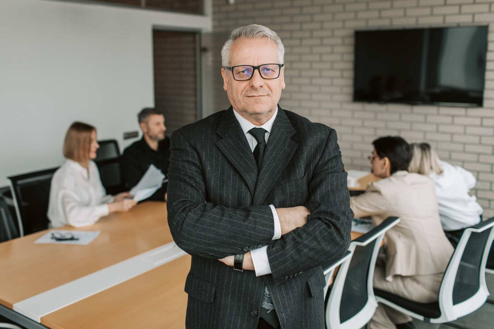 A senior man in a pinstripe suit stands with arms crossed in a bright boardroom while colleagues confer at a table behind him