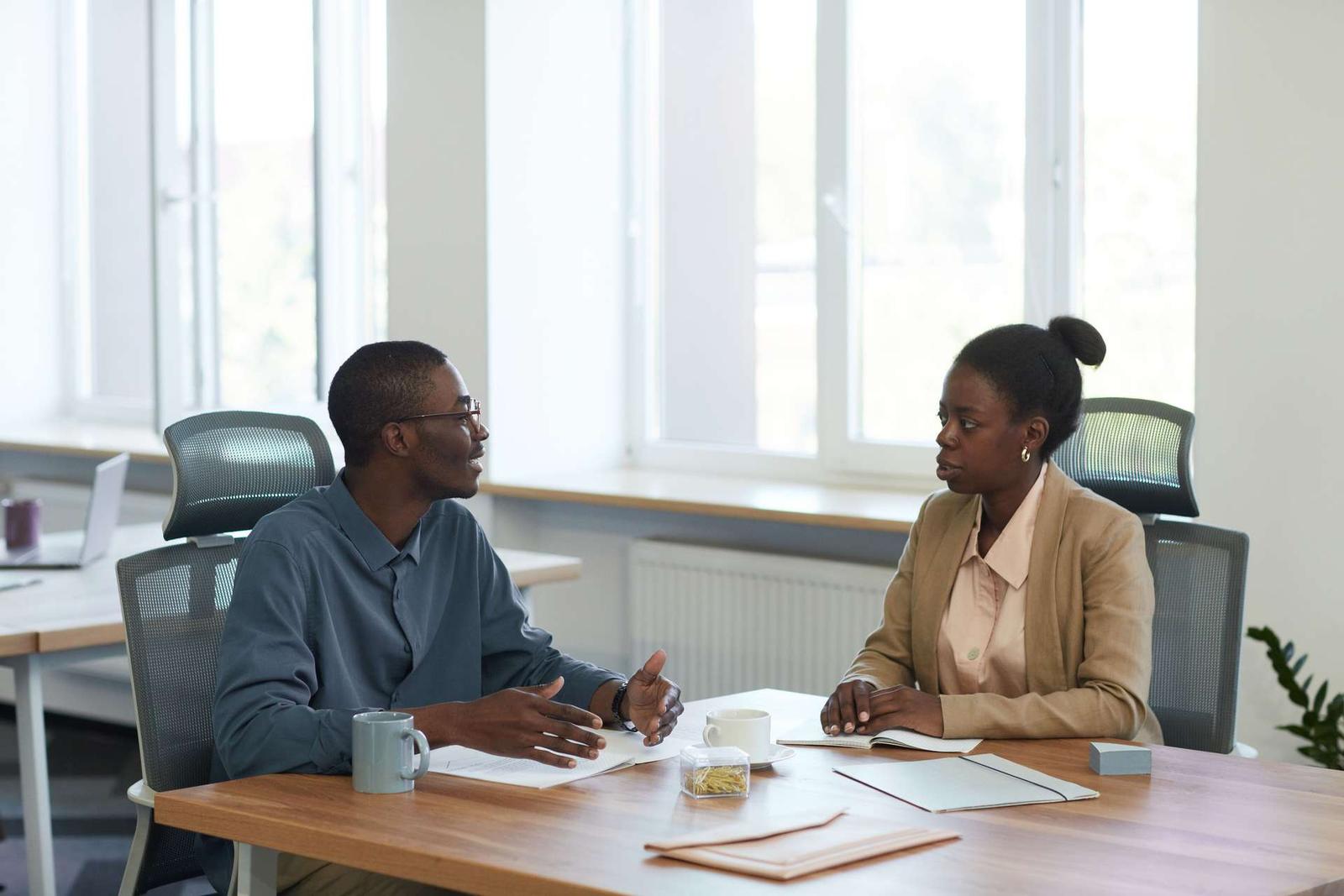 A man in a grey shirt gestures while talking with a woman in a tan blazer across a desk with open notebooks and coffee mugs, in a bright office with large windows