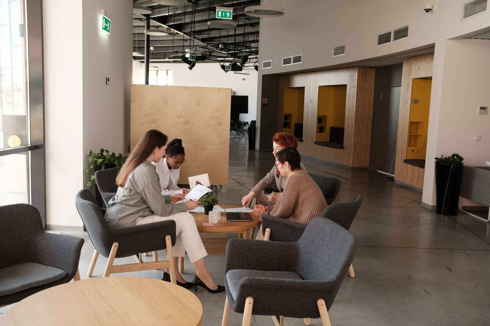 Four women in business dress review papers together at a coffee table in the lounge of a bright, high-ceilinged hall