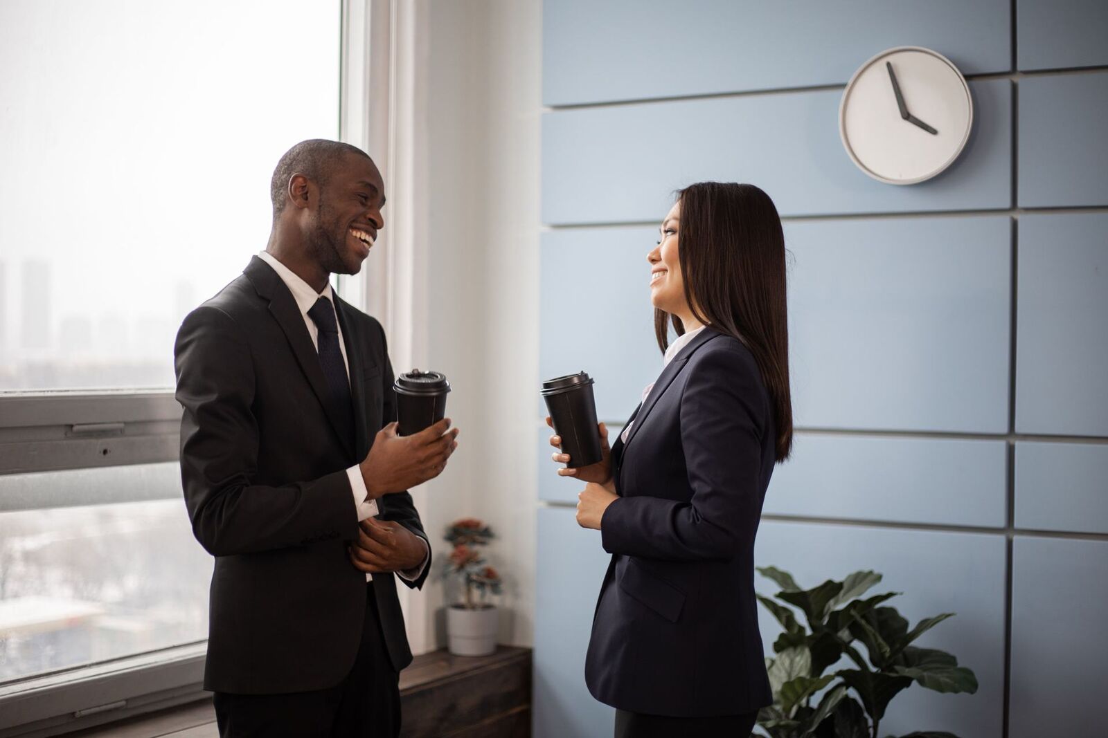 A man and a woman in dark business suits stand laughing together near a bright window, each holding a to-go coffee cup, with a round wall clock on the wall behind them