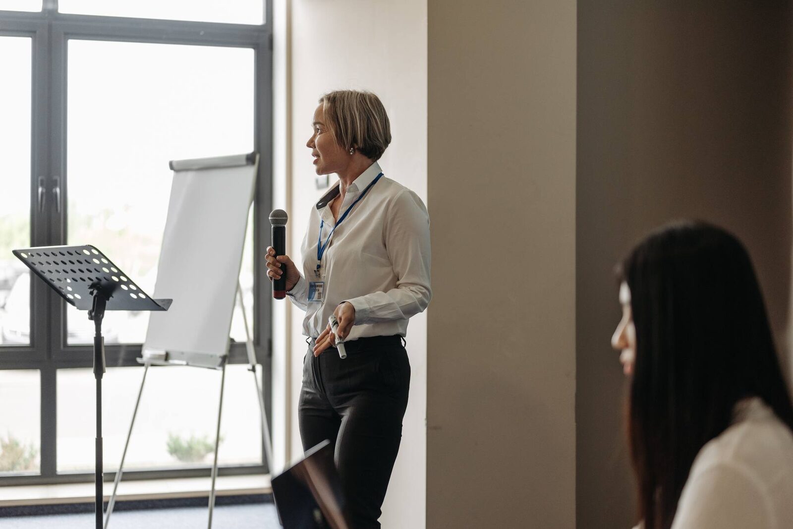 A woman with a conference lanyard speaks into a handheld microphone beside a flipchart and a music-stand lectern, tall bright windows behind her