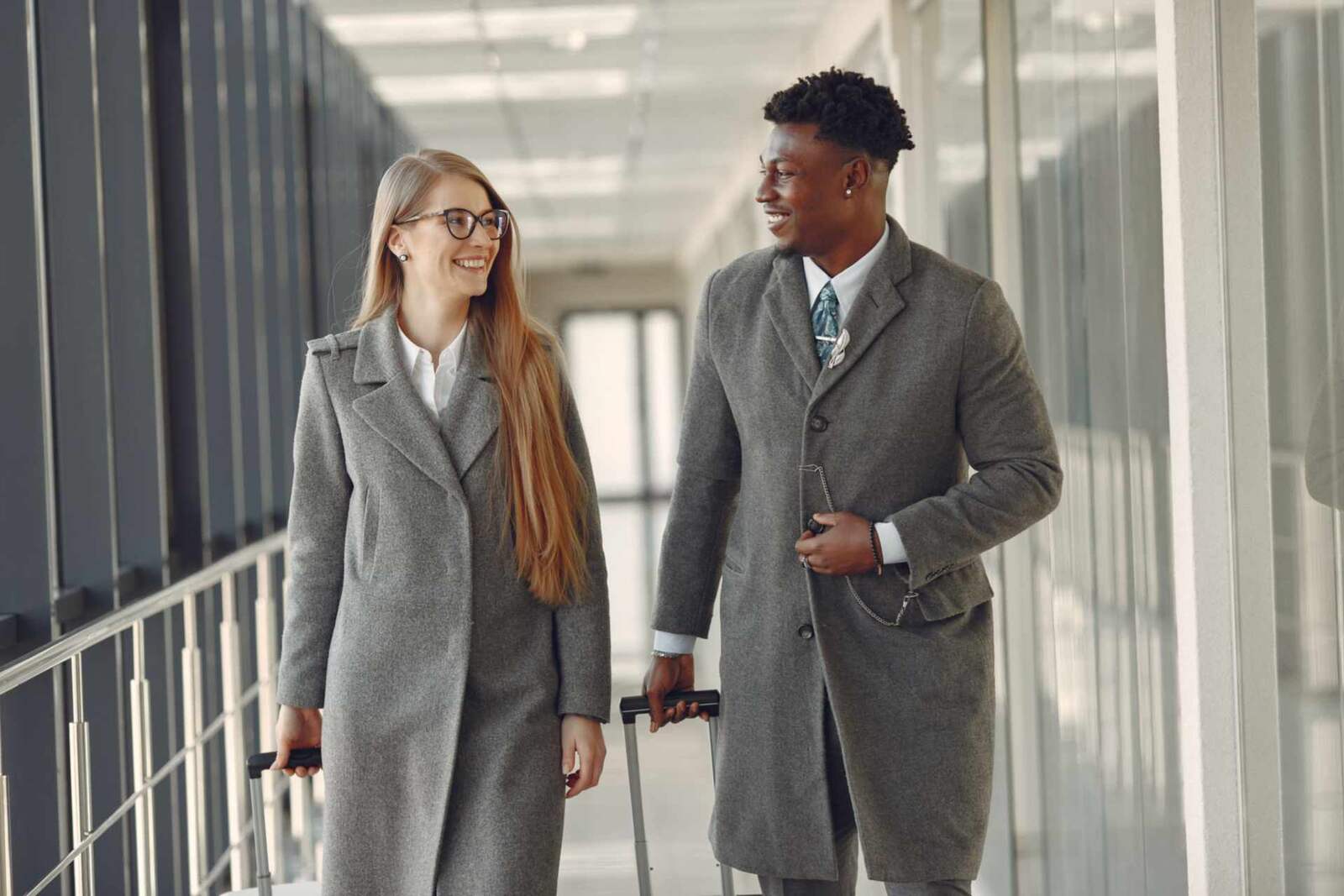 A smiling woman and man in long grey coats pull rolling cases together along a bright glass skybridge