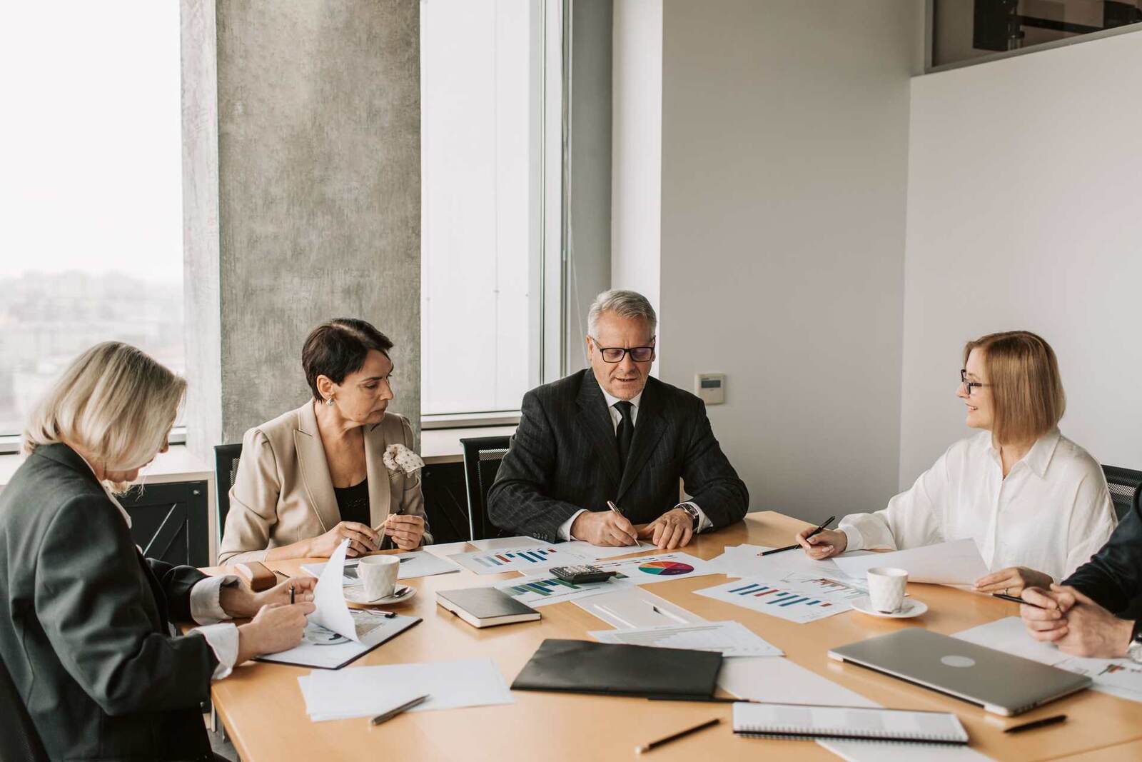 Five senior delegates in suits and blazers work through printed charts together at a bright meeting table by a window