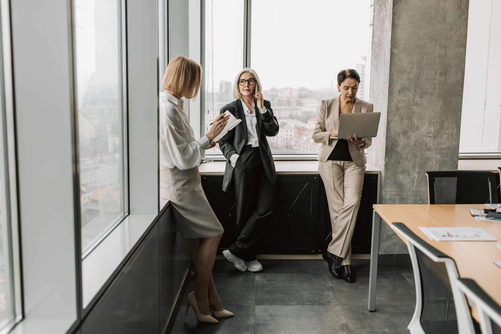 Three senior women in business suits by floor-to-ceiling windows above a city skyline, one taking notes, one on the phone, one working on a laptop