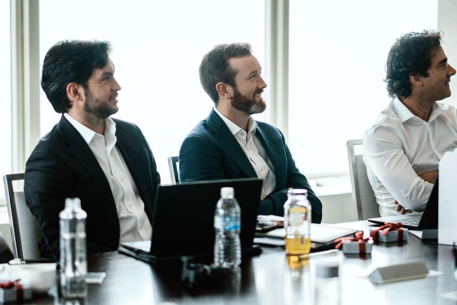 Three men in business attire sit at a table with laptops open and small ribbon-tied gift boxes, smiling as they listen to an unseen speaker in a bright, window-lit room