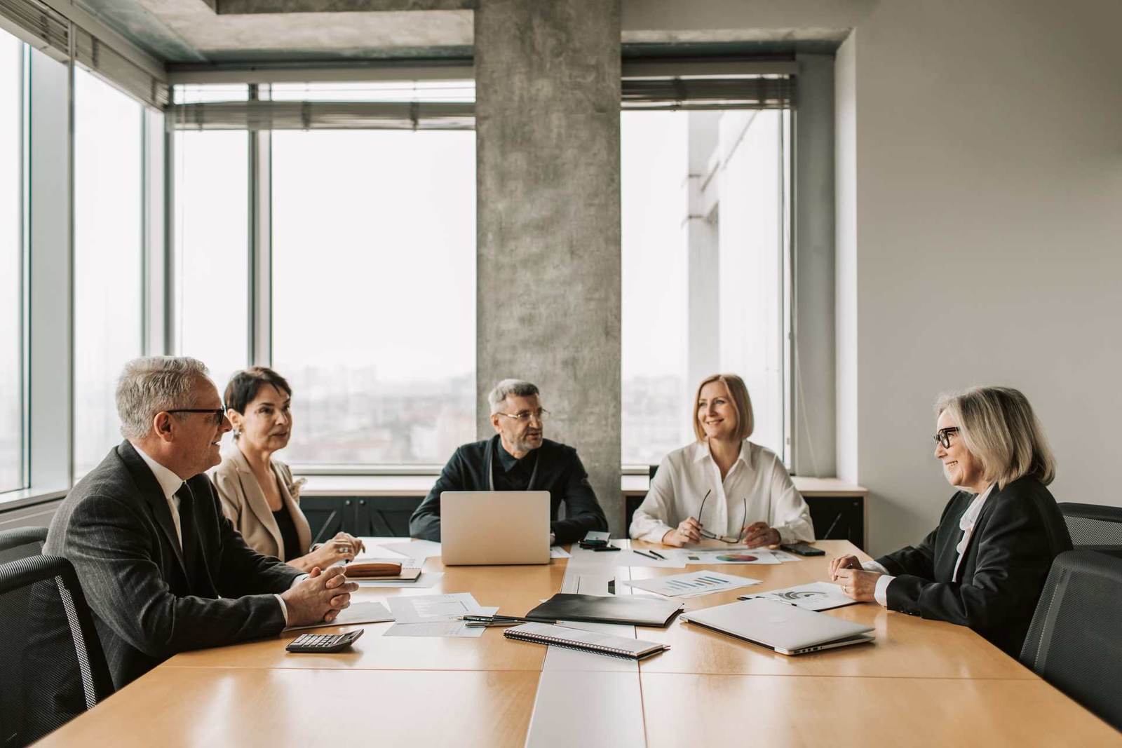 Five colleagues in business attire around a boardroom table with laptops and printed charts, city skyline visible through floor-to-ceiling windows