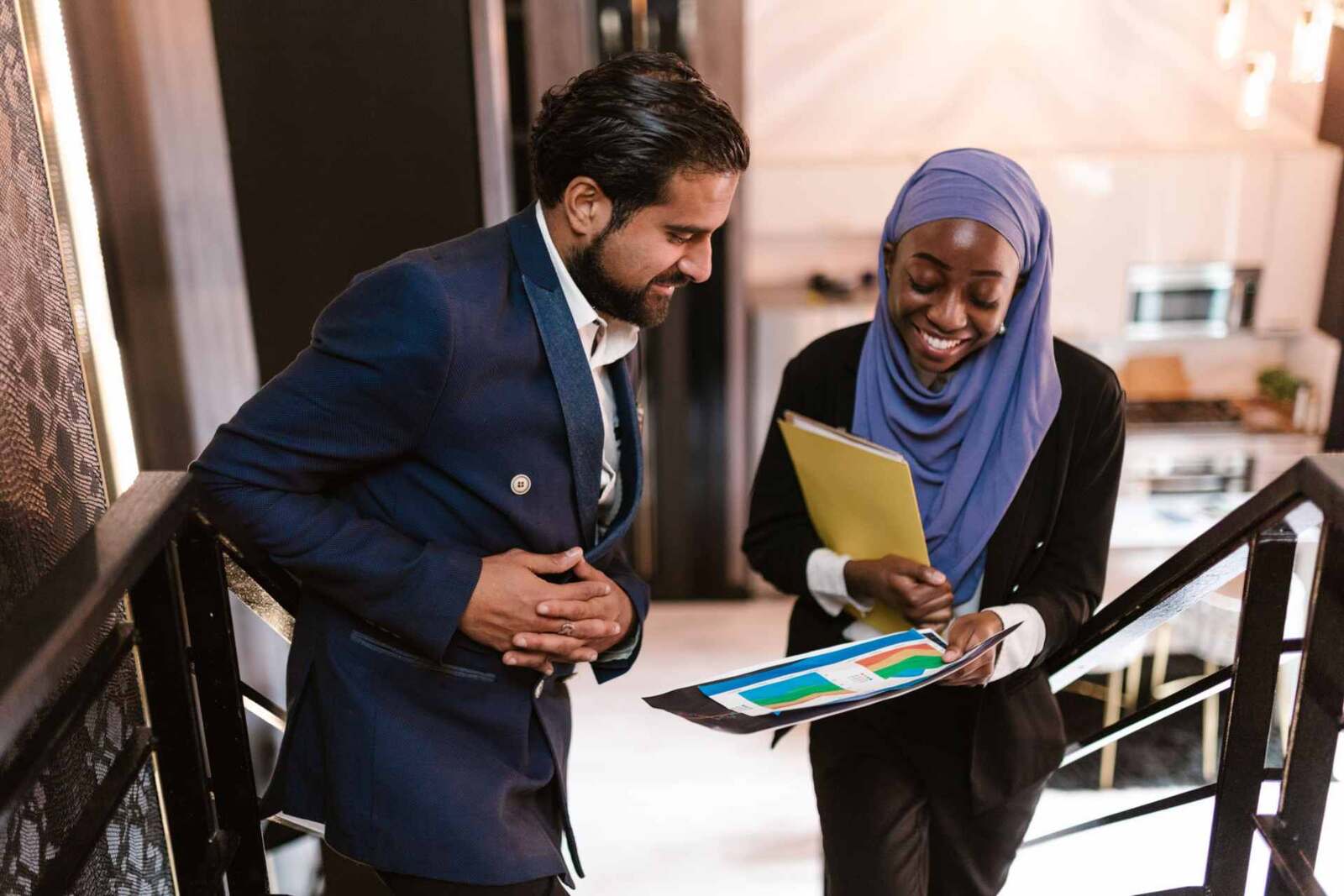 Two colleagues in business attire look over a printed chart together while standing on a staircase in a bright venue