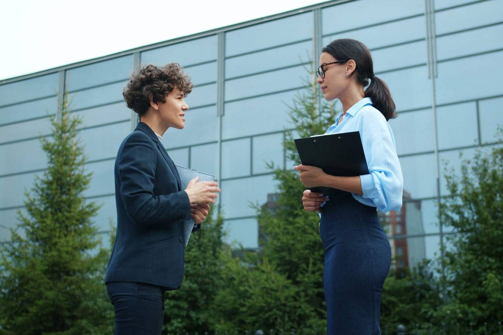 Two women in business dress talk outside a glass-fronted office building, one holding a clipboard, under an overcast bright sky