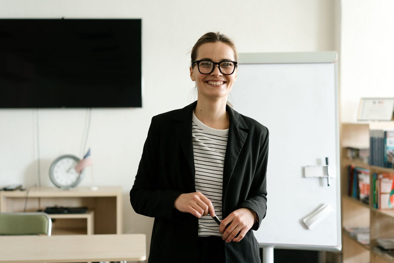 A smiling woman in a black blazer stands in front of a blank flipchart, marker in hand, as if about to begin a talk