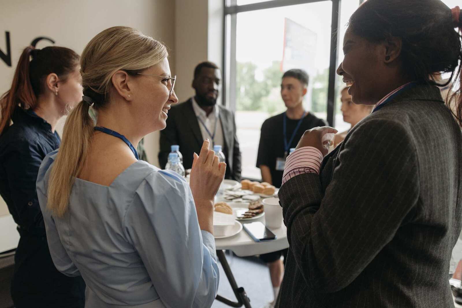 Two women with conference lanyards laugh mid-conversation by a table of water and pastries, colleagues talking behind them at a bright window