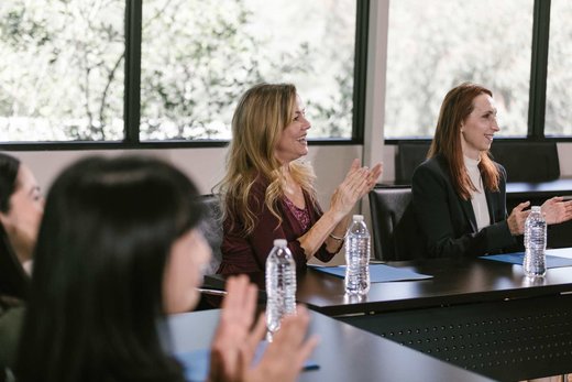 Two women applaud from their seats at a conference table lined with water bottles and folders, bright windows behind them