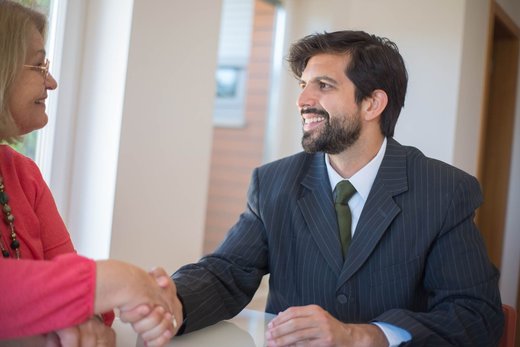 A man in a pinstripe suit smiles and shakes hands with an older woman across a table, in a bright room with tall windows