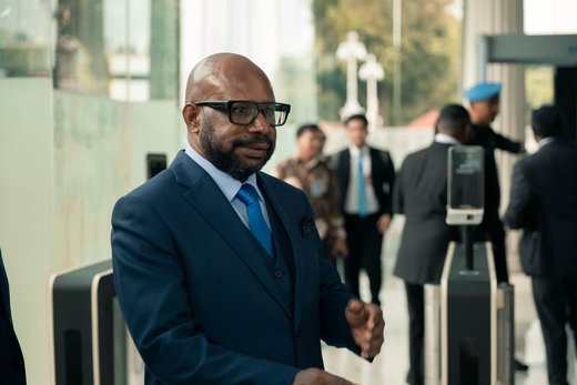 A bearded man in a navy three-piece suit and glasses walks through a bright lobby past a walk-through security scanner, with uniformed personnel and other visitors visible in the background