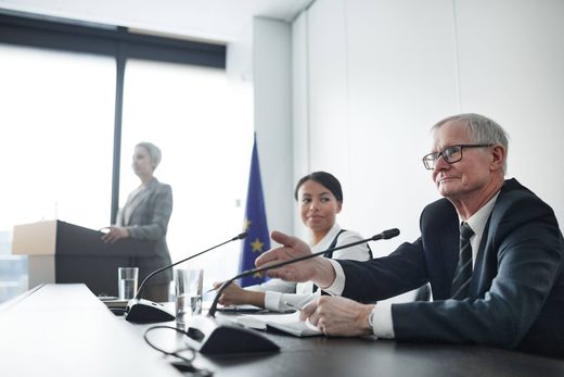 An EU flag stands behind a panel of three speakers at microphones in a bright, formal conference room