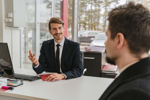 A man in a navy suit gestures while talking across a white desk to a colleague, in a bright glazed office