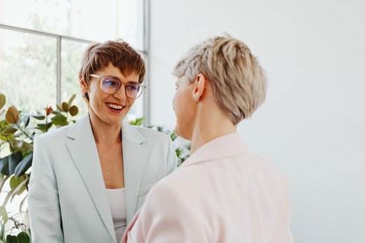 Two women in pale blazers laugh mid-conversation beside a bright window and potted plants