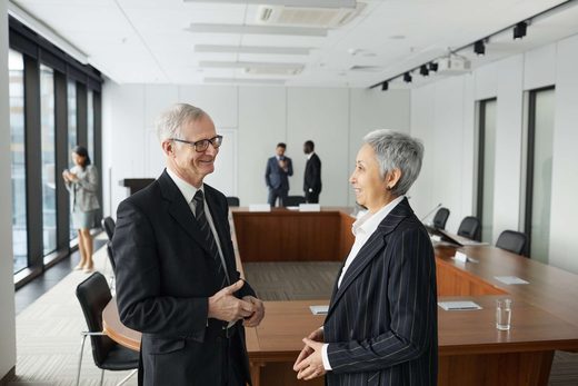 Two senior delegates chat and smile in profile beside an empty U-shaped hearing-room table before the room fills, floor-to-ceiling windows and two colleagues in soft focus behind them