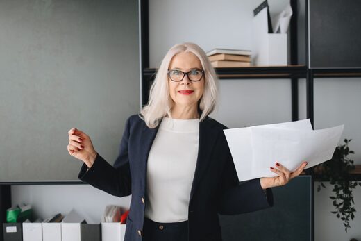 A woman in a navy blazer holds a sheaf of papers mid-gesture in a bright office lined with shelves