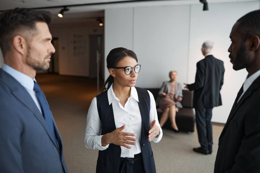Three colleagues in business attire talk in a bright office corridor, a woman gesturing mid-explanation between two men, with two more colleagues in conversation in the background