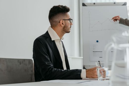 A man in glasses and a black blazer sits at a conference table, attentively watching an off-frame hand point to a hand-drawn line chart on a flipchart in a bright white room