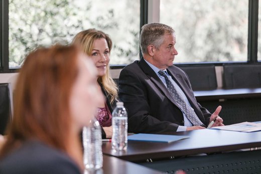 A woman and a man listen attentively at a conference table lined with water bottles, a colleague blurred in the foreground and trees visible through the windows behind them