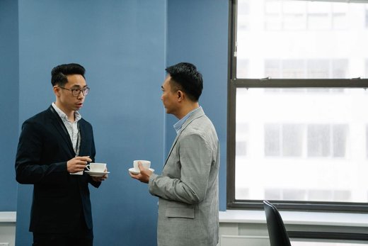 Two colleagues in blazers stand talking over cups of coffee near a large window in a blue-walled meeting room, one wearing a lanyard badge